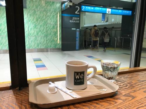 We CAFFE エキュート大宮店のカウンターテーブルに置いたホットコーヒーと水、窓の向こうに見える京浜東北線の標識