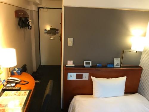 アパホテル金沢駅前のシングルルームの部屋の机とベッドと壁にかかるハンガーと部屋の入口